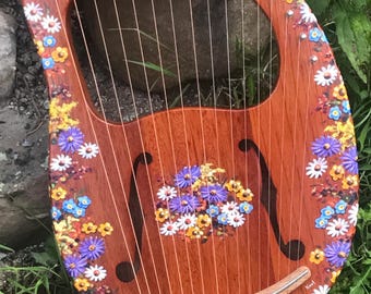 May include: A wooden lyre with a floral design painted on the body. The lyre has 10 strings and a gold-coloured tuning key. The lyre is sitting on a bed of green grass.