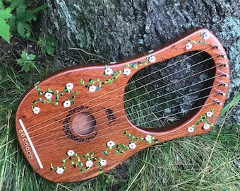Può includere: Un liuto di legno con un disegno floreale dipinto sul corpo. Il liuto ha fiori bianchi e viti verdi dipinti su uno sfondo marrone. Il liuto ha un foro sonoro al centro e una fila di piroli in cima.