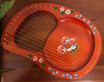 May include: A wooden lyre with a red finish and painted floral and mushroom designs. The lyre has 10 strings and a soundboard with a rounded shape.