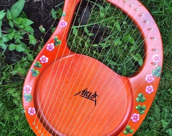 May include: A wooden lyre with a floral design painted on the side. The lyre has 15 strings and is resting on a bed of green grass. The lyre is signed "Miklot".