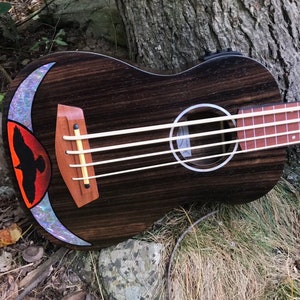 Op de afbeelding: Een donkerhouten ukulelebas met een zwarte vogelinleg op het klankgat. De inleg is omgeven door een parelmoerachtige maanvorm.