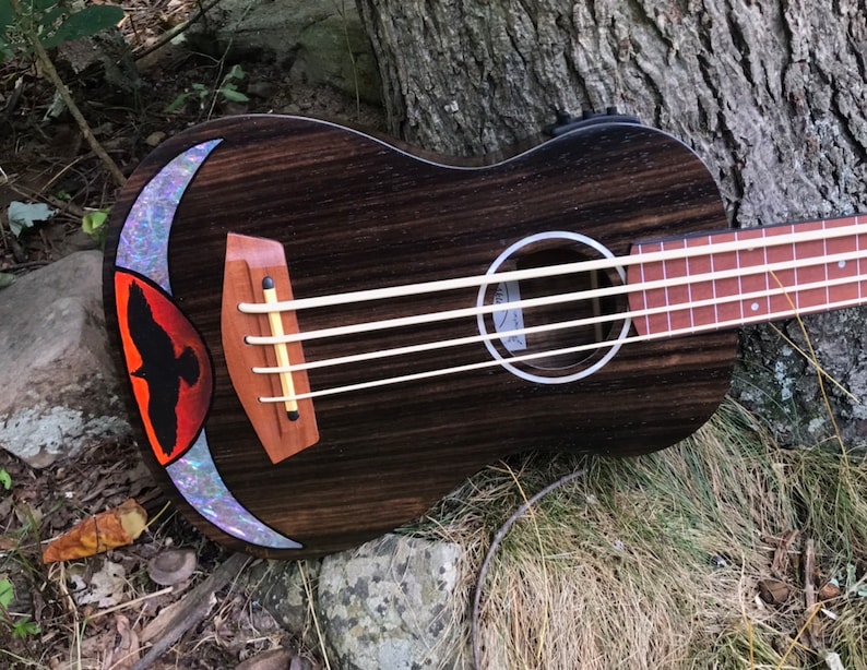Op de afbeelding: Een donkerhouten ukulelebas met een zwarte vogelinleg op het klankgat. De inleg is omgeven door een parelmoerachtige maanvorm.