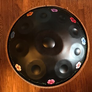 Op de afbeelding: Een zwarte stalen handpan met een bruine touwrand en geschilderde bloemen in verschillende kleuren rond de rand.