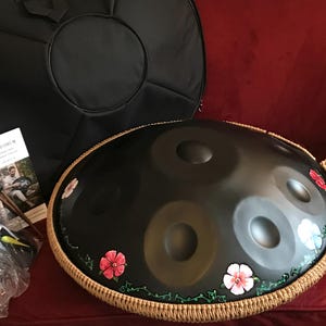 Op de afbeelding: Een zwarte handpan met een bruine touwrand en geschilderde bloemen. De handpan ligt op een rood oppervlak met een zwarte draagtas en een standaard ernaast.