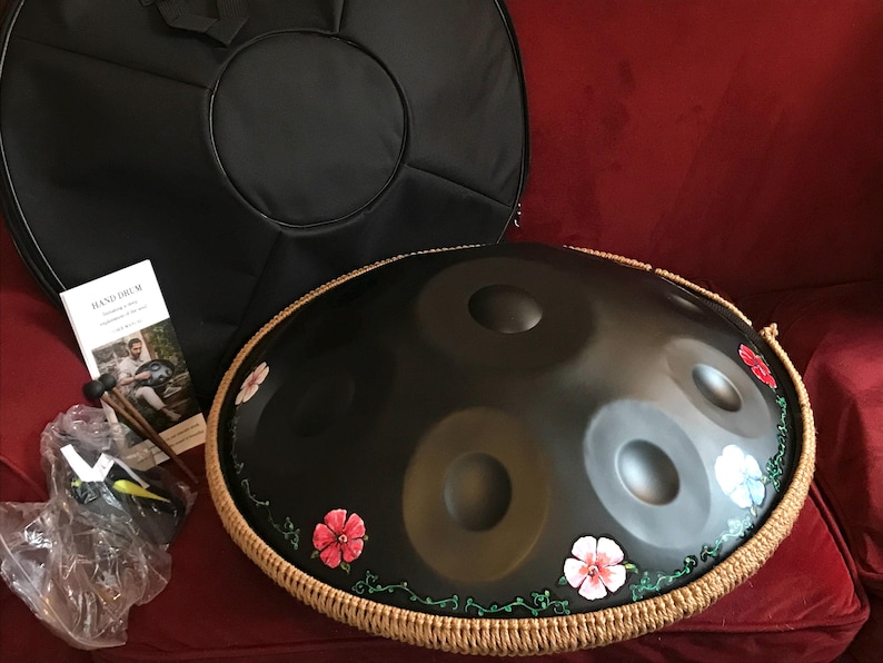 Op de afbeelding: Een zwarte handpan met een bruine touwrand en geschilderde bloemen. De handpan ligt op een rood oppervlak met een zwarte draagtas en een standaard ernaast.
