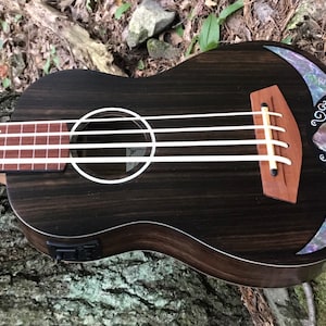 K&ouml;nnte beinhalten: Eine schwarze Ukulele mit Holzmaserung und einer Perlmutt-Einlage um das Schallloch. Die Ukulele hat vier Saiten und eine Muscheleinlage am oberen Korpus.