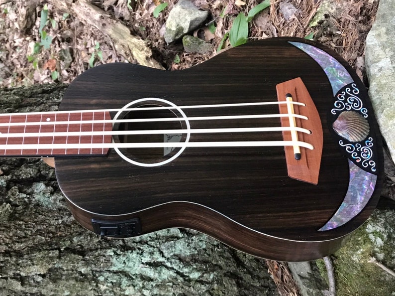 K&ouml;nnte beinhalten: Eine schwarze Ukulele mit Holzmaserung und einer Perlmutt-Einlage um das Schallloch. Die Ukulele hat vier Saiten und eine Muscheleinlage am oberen Korpus.