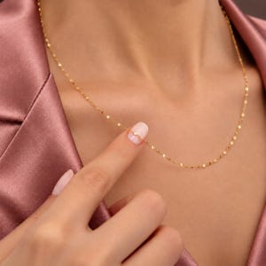 May include: A delicate gold chain necklace featuring small, flat, reflective links that catch the light. The necklace rests against the collarbone, showcasing a thin, minimalist design with a polished finish that creates a subtle, shimmering effect.