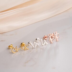 May include: A collection of stud earrings in gold, silver, and rose gold. Each earring features a different letter of the alphabet. The earrings are displayed on a white surface with a soft, neutral-colored fabric in the background.