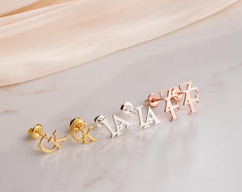 May include: A selection of stud earrings in gold, silver, and rose gold. Each earring displays a different letter of the alphabet. The earrings are presented on a white surface with a soft, neutral-coloured fabric in the background.