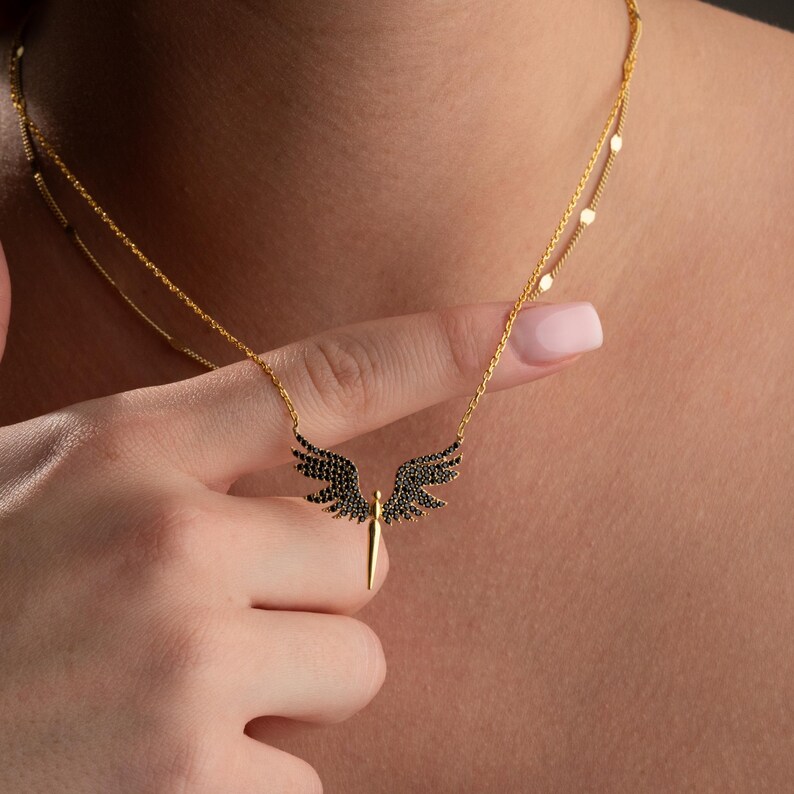 May include: A gold-colored necklace with a pendant featuring black wings and a central gold-colored sword. The necklace has a delicate chain design and is displayed against a skin-toned background.