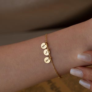 May include: Gold chain bracelet with three small round charms, each with a letter engraved on it. The letters are R, T, and B.