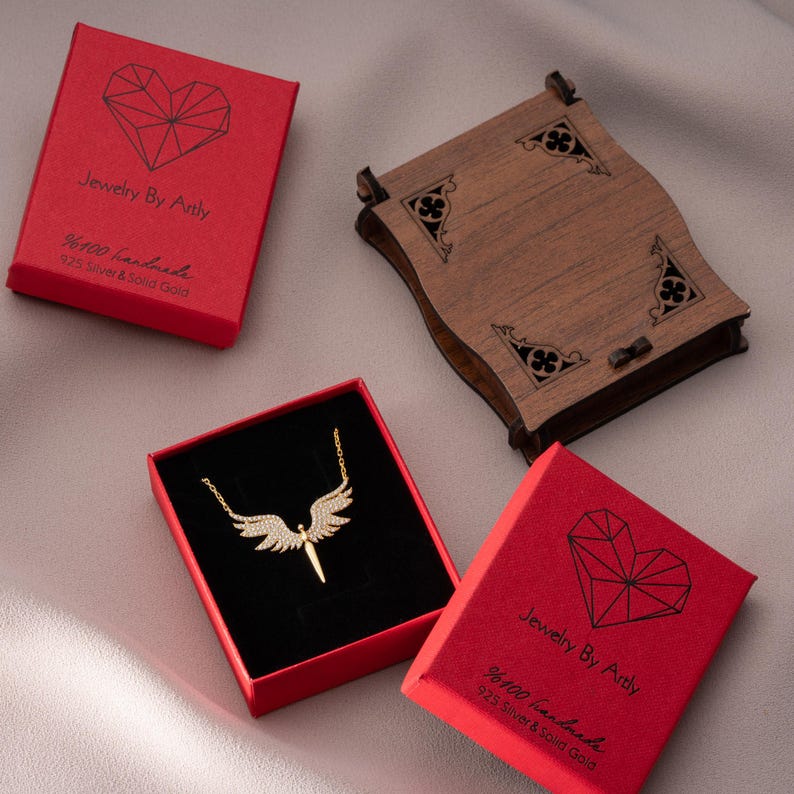 May include: A gold-colored necklace with an angel wing design, displayed in a red box with black velvet lining. The box is next to two other red boxes and a wooden box. The text on the red boxes reads "Jewelry By Artly" and "%100 handmade 925 Silver & Solid Gold."