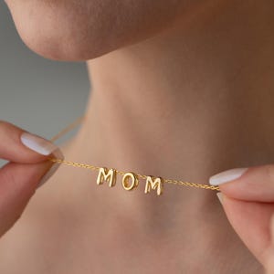 Könnte beinhalten: Eine goldene Halskette mit dem Wort "MOM" in einzelnen Buchstaben.