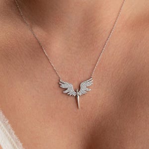 May include: A silver-toned necklace featuring a pendant shaped like angel wings. The wings are encrusted with small, sparkling stones, and the pendant has a central, pointed element. The necklace is displayed against a neutral background.