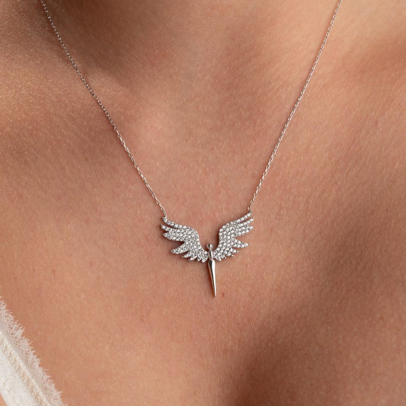 May include: A silver-toned necklace featuring a pendant shaped like angel wings. The wings are encrusted with small, sparkling stones, and the pendant has a central, pointed element. The necklace is displayed against a neutral background.
