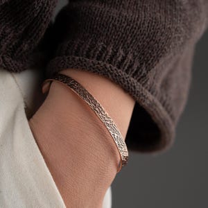 May include: A copper-toned metal cuff bracelet featuring a textured, irregular surface pattern of raised lines and grooves. The bracelet has an open-ended design and a metallic finish that catches the light, showcasing a detailed, organic relief texture.