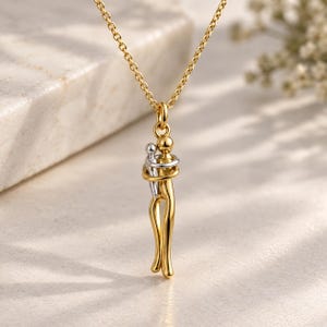 May include: A gold-toned necklace featuring a pendant of two stylized figures in an embrace. The larger figure is gold-toned, while the smaller figure is silver-toned. The pendant hangs from a delicate gold-toned chain with a simple link design.
