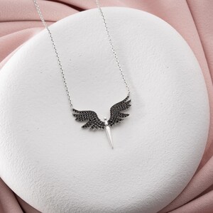 May include: A silver necklace featuring a pendant shaped like angel wings. The wings are detailed with black gemstones, and the pendant has a central, pointed element. The necklace is displayed on a white surface with a pink background.