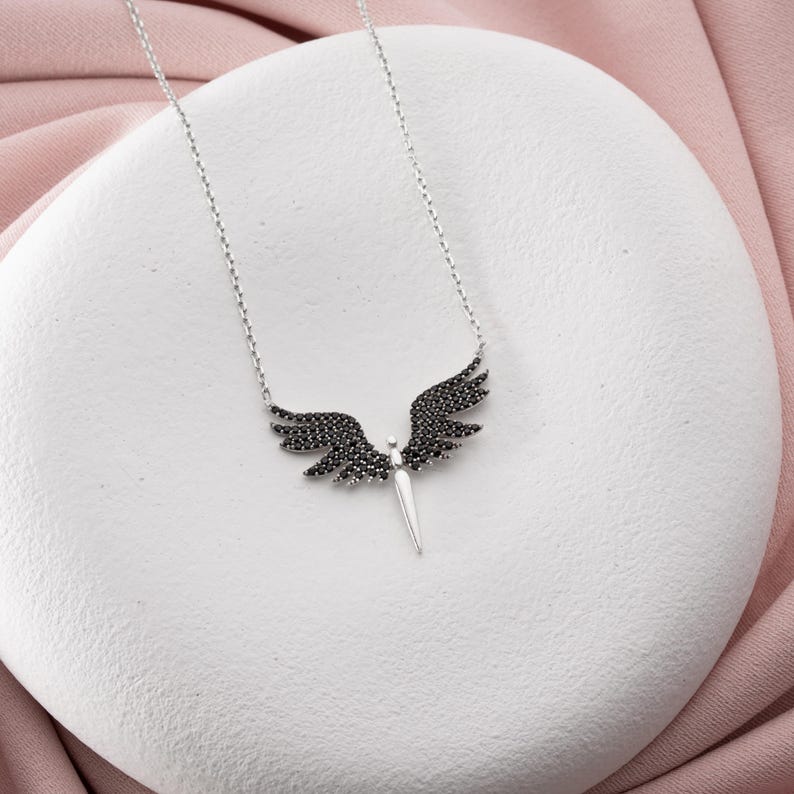 May include: A silver necklace featuring a pendant shaped like angel wings. The wings are detailed with black gemstones, and the pendant has a central, pointed element. The necklace is displayed on a white surface with a pink background.