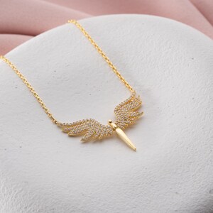 May include: A gold-colored necklace featuring a pendant with a wing design. The wings are adorned with small, sparkling stones. The pendant has a central, elongated shape. The necklace is displayed on a white surface.