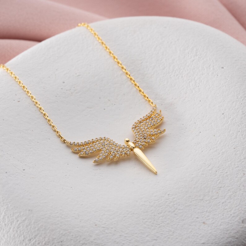 May include: A gold-colored necklace featuring a pendant with a wing design. The wings are adorned with small, sparkling stones. The pendant has a central, elongated shape. The necklace is displayed on a white surface.