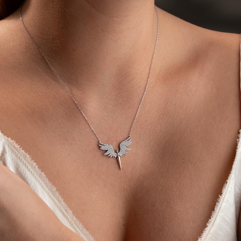 May include: A silver necklace featuring a pendant shaped like angel wings. The wings are encrusted with small, sparkling stones. The necklace is displayed on a person's chest, showcasing the delicate design and craftsmanship of the jewelry.