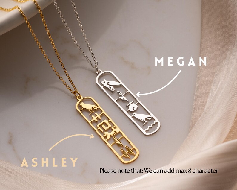 May include: Two necklaces with pendant designs. One is gold-colored, the other is silver. Each pendant has a rectangular shape with cut-out bird and hieroglyphic designs. The gold necklace is labeled "ASHLEY", and the silver necklace is labeled "MEGAN".