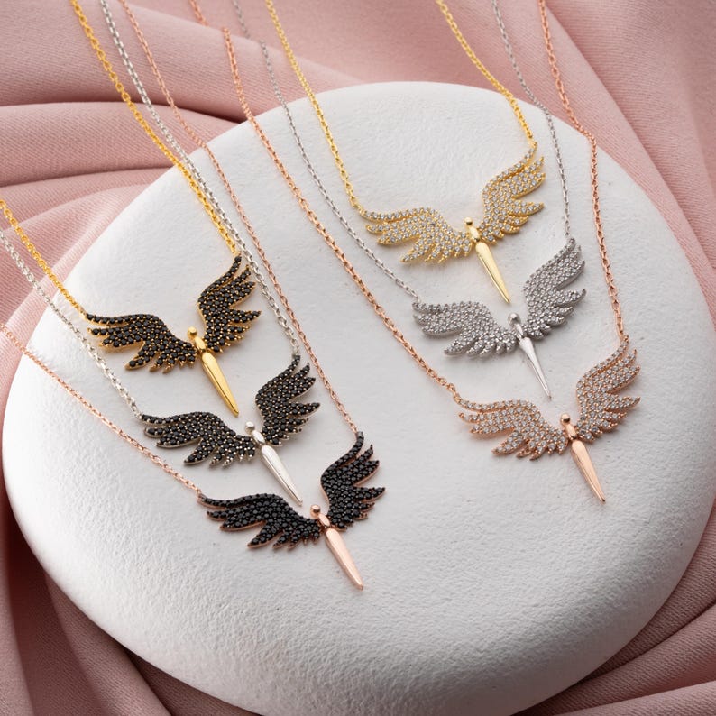 May include: A collection of angel wing necklaces in gold, silver, and rose gold tones. Each necklace features a pendant with detailed wings and a central body. The wings are adorned with black or clear stones.