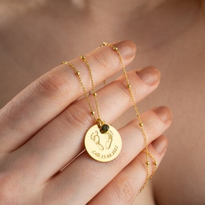 May include: A gold-toned necklace featuring a delicate chain with small beads and a circular pendant. The pendant is engraved with two baby footprints and the text Lilli 15.08.2021, accented by a small, round, dark green gemstone charm.