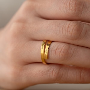 Peut inclure: Une bague en or avec les noms "Emma" et "Cass" gravés dessus. La bague est portée au doigt d'une personne.