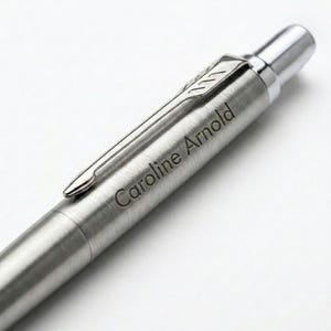 May include: A brushed silver ballpoint pen featuring a polished chrome pocket clip and a click-action mechanism at the top. The barrel of the pen is engraved with the name Caroline Arnold in a clean, dark, sans-serif font, creating a metallic finish.