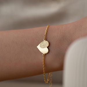 May include: A gold chain bracelet with a double heart charm. The larger heart has a fingerprint design engraved on it.