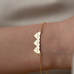 Op de afbeelding: Een goudkleurige armband met drie hartvormige bedels. Elke bedel is gegraveerd met een naam: Emma, James en Stella. De armband is delicaat en lijkt van een fijne ketting te zijn gemaakt.