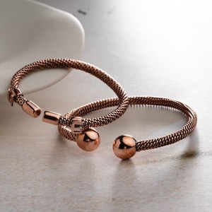 May include: Two copper-toned cuff bracelets featuring a textured, woven-style band. Each bracelet is open-ended and finished with a smooth, polished spherical bead at each terminal end, providing a consistent metallic appearance throughout.