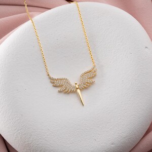 May include: A gold-colored necklace with a pendant shaped like angel wings. The wings are encrusted with small, clear gemstones. The pendant is suspended from a delicate chain. The necklace is displayed on a white surface.