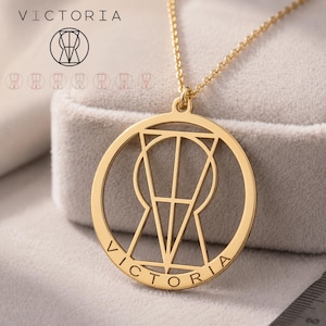 May include: A gold-toned circular pendant necklace featuring a geometric cutout design of the letter V and the name VICTORIA engraved along the bottom edge. The pendant hangs from a delicate gold-toned chain, showcasing a polished metallic finish.