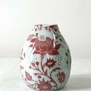 May include: A small, ceramic vase with a light blue background and a red floral pattern. The vase has a glossy finish and a rounded shape, perfect for displaying flowers or as a decorative piece. The vase is approximately 6 inches tall.
