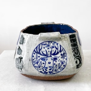 May include: A handcrafted ceramic bowl with a textured white exterior, featuring blue and black decorative patterns. The bowl has a brown base and a deep blue interior. The design includes sheep and a deer with floral elements.