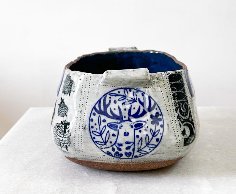 May include: A handcrafted ceramic bowl with a textured white exterior, featuring blue and black decorative patterns. The bowl has a brown base and a deep blue interior. The design includes sheep and a deer with floral elements.