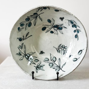 May include: A round ceramic bowl with a white glazed finish, featuring hand-painted blue olive branches and small black bees scattered across the interior surface. The bowl has a textured, slightly uneven rim and a deep, wide basin design.
