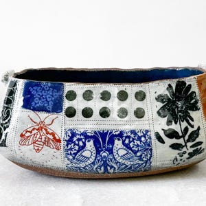 May include: Handmade ceramic bowl with a patchwork design. The bowl features a dark blue interior and a brown base. The exterior is decorated with various patterns, including a red moth, blue floral motifs, and black flower illustrations. The bowl has two small handles.