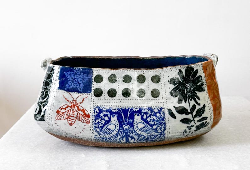 May include: Handmade ceramic bowl with a patchwork design. The bowl features a dark blue interior and a brown base. The exterior is decorated with various patterns, including a red moth, blue floral motifs, and black flower illustrations. The bowl has two small handles.