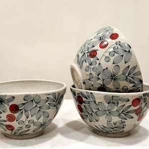 May include: Three ceramic bowls with a speckled off-white base featuring a black line-art pattern of leaves and flowers accented with small, solid red circular berries. One bowl sits to the left, while two are stacked on the right side.
