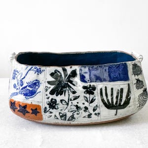May include: Handmade ceramic bowl with a patchwork design. The bowl features a dark blue interior and is decorated with blue birds, black flowers, and sheep on a white background. The rim has two small handles.