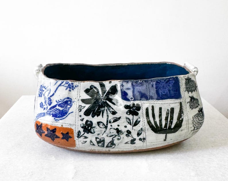 May include: Handmade ceramic bowl with a patchwork design. The bowl features a dark blue interior and is decorated with blue birds, black flowers, and sheep on a white background. The rim has two small handles.