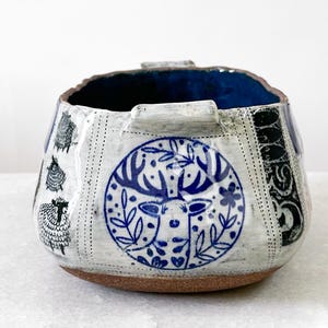 May include: A ceramic bowl with a blue interior and a textured exterior. The bowl features a deer design in a blue circle, sheep illustrations, and black decorative patterns. The base is a warm brown color.