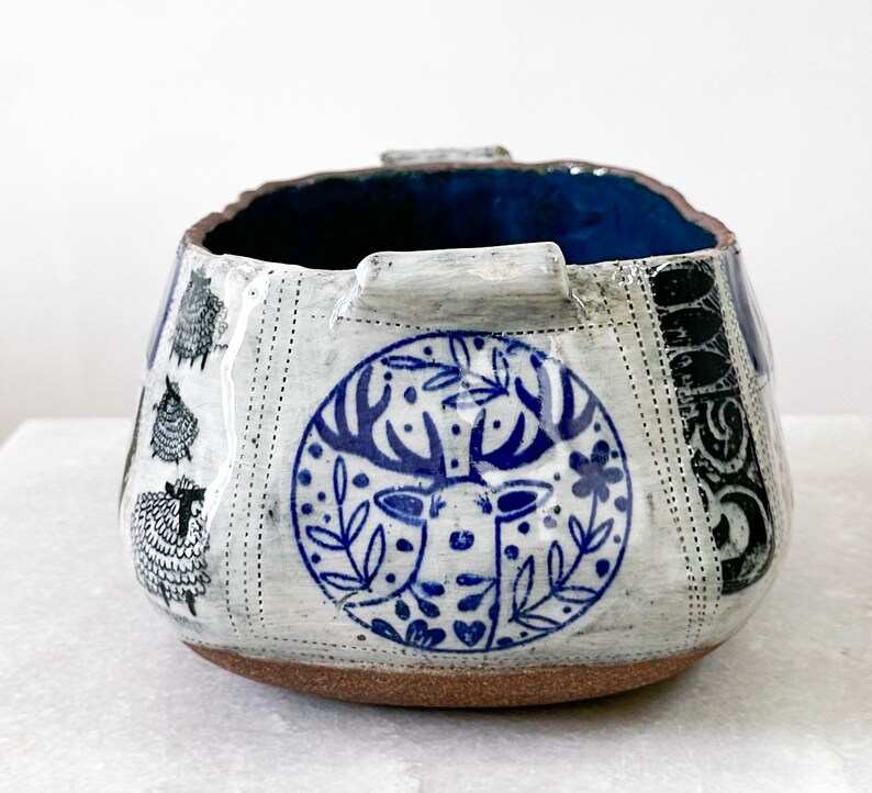 May include: A ceramic bowl with a blue interior and a textured exterior. The bowl features a deer design in a blue circle, sheep illustrations, and black decorative patterns. The base is a warm brown color.