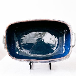 May include: A rectangular ceramic baking dish with a deep blue interior and a lighter blue rim. The dish has a rough, textured exterior with a brown edge and two handles. It sits on a black metal stand, perfect for serving.