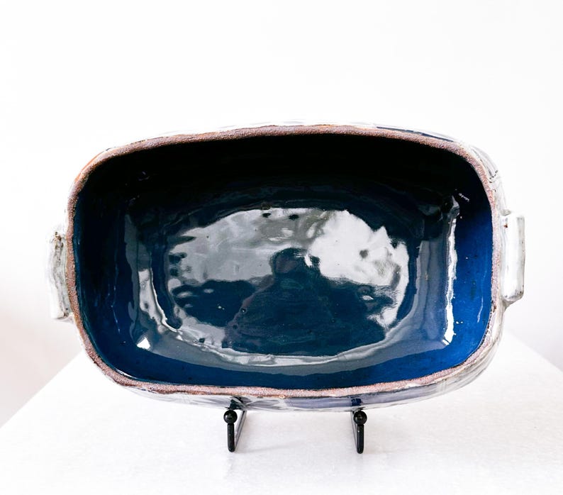 May include: A rectangular ceramic baking dish with a deep blue interior and a lighter blue rim. The dish has a rough, textured exterior with a brown edge and two handles. It sits on a black metal stand, perfect for serving.